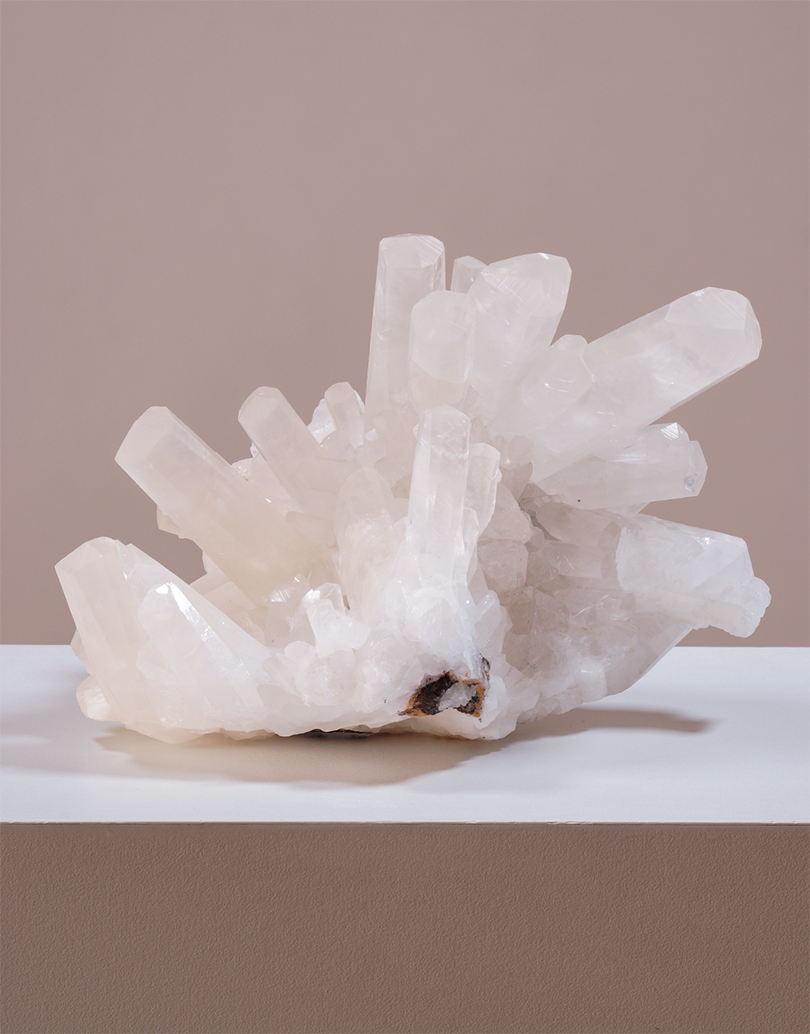A natural white crystal cluster with multiple translucent pointed mineral stalks, displayed on a white platform against a warm beige background.