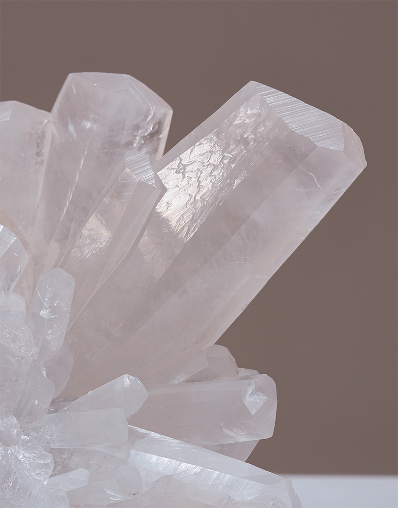 A close-up studio shot of a translucent white crystal cluster, including a prominent crystal point, set against a solid warm taupe-brown background.